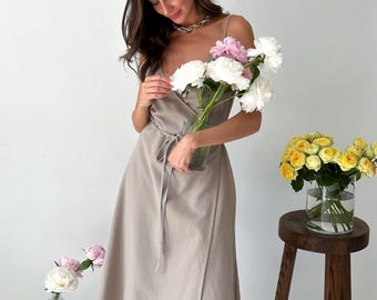 May include: A woman wearing a beige wrap dress and holding a bouquet of white and pink peonies. She is standing in front of a white wall.