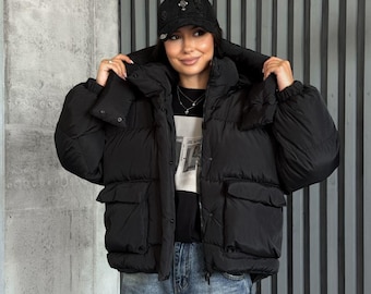May include: A black puffer jacket with a high collar, front snap buttons, and two large flap pockets. The jacket is worn over a black t-shirt with white text and blue denim jeans. A black baseball cap with a small silver emblem is also worn.