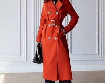 May include: A vibrant orange double-breasted trench coat featuring a wide collar, shoulder epaulets, and a matching belt with a circular buckle. The coat is worn over a dark brown shirt and dark brown wide-leg trousers, paired with a black handbag.