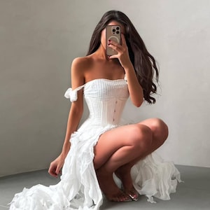 Op de afbeelding: Een witte off-shoulder jurk met een textuurrijk, gesmokt lijfje met een centraal knoopdetail en een volumineuze, geplooide rok met een asymmetrische zoom. Het kledingstuk heeft korte, gedrapeerde mouwen en een getailleerde korset-stijl.