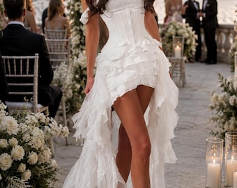 May include: A white strapless dress featuring a corset-style bodice with a smocked texture and a high-low skirt layered with multiple cascading ruffles. The garment is worn with clear high-heeled sandals, showcasing a layered, voluminous silhouette.
