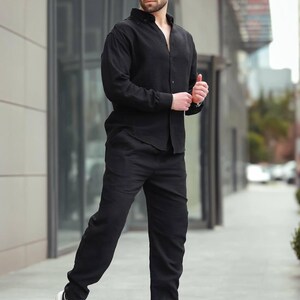May include: A black linen shirt and trousers outfit. The model is wearing sunglasses and white trainers. The ensemble is suitable for casual or semi-formal wear. The outfit is all black.