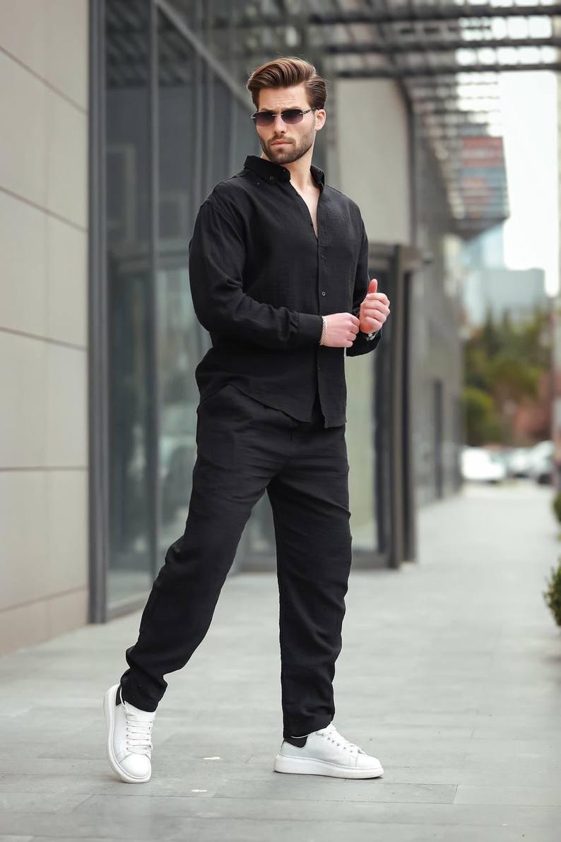 May include: A black linen shirt and trousers outfit. The model is wearing sunglasses and white trainers. The ensemble is suitable for casual or semi-formal wear. The outfit is all black.