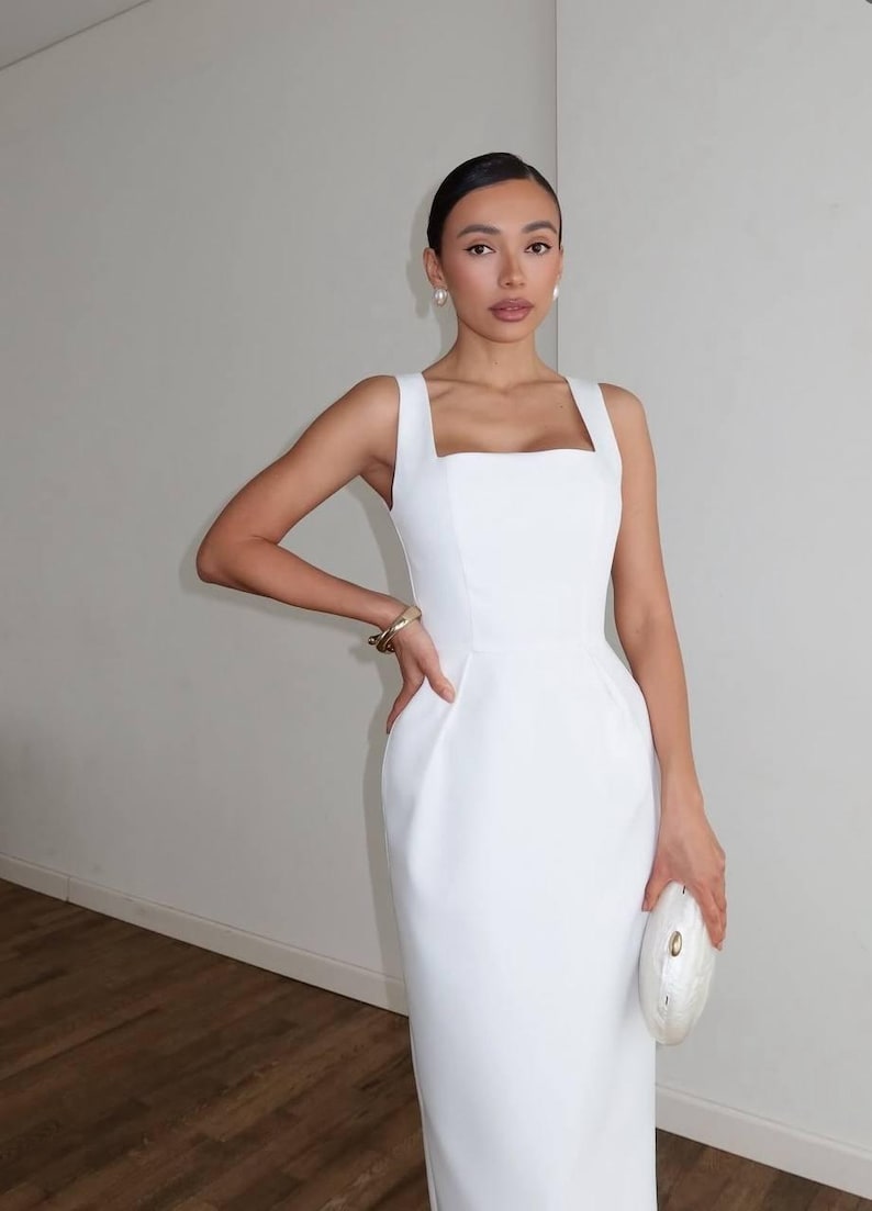 K&ouml;nnte beinhalten: Ein wei&szlig;es Kleid mit quadratischem Ausschnitt, eng anliegendem Oberteil und geradem Rock. Das Kleid hat ein schlichtes, elegantes Design, ideal f&uuml;r formelle Anl&auml;sse. Das Model tr&auml;gt ein goldenes Armband, Perlenohrringe und eine wei&szlig;e Clutch.