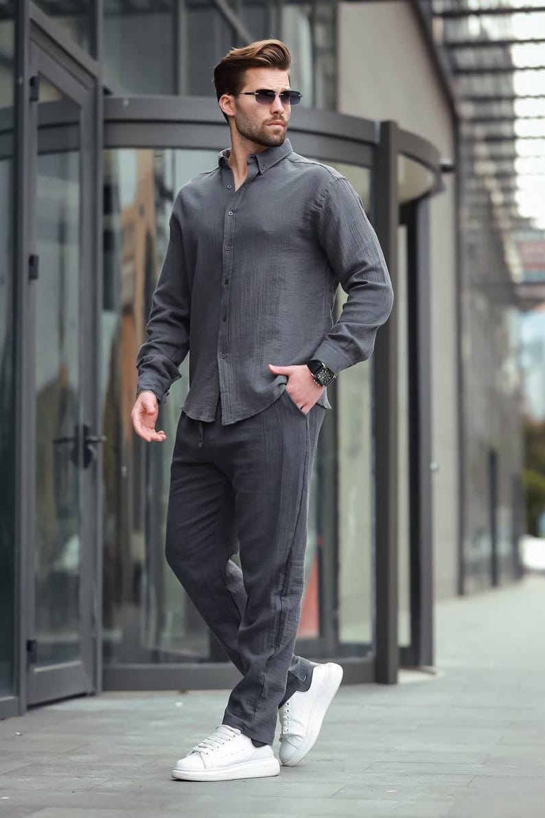 May include: A person in a charcoal grey linen shirt and trousers, paired with white trainers and sunglasses. The long-sleeved shirt is buttoned, and the trousers have a relaxed fit. The outfit suggests a casual, contemporary style, suitable for warmer weather.