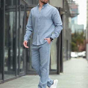 May include: A light blue linen shirt and matching trousers set. The long-sleeved shirt is buttoned down and the trousers are straight-legged. The outfit is paired with white trainers and sunglasses. The person is wearing a black watch.