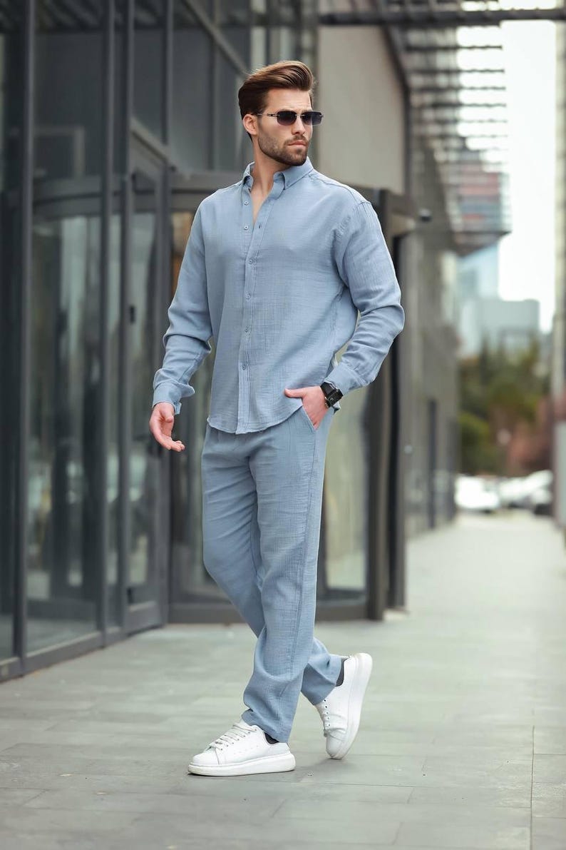 May include: A light blue linen shirt and matching trousers set. The long-sleeved shirt is buttoned down and the trousers are straight-legged. The outfit is paired with white trainers and sunglasses. The person is wearing a black watch.
