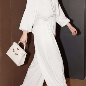 May include: A white outfit comprising a long-sleeved blouse and wide-leg trousers. A white handbag with gold-coloured hardware is held. The outfit is completed with white pointed-toe heels. The background features a brown and black wall.