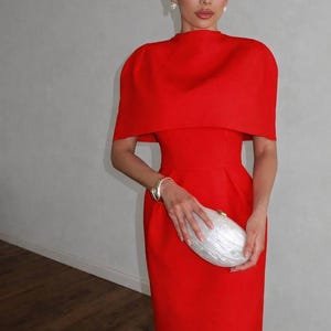 K&ouml;nnte beinhalten: Ein leuchtend rotes Kleid mit einem passenden, kapuzenartigen Oberteil. Das Kleid wird mit einer wei&szlig;en Clutch und einem goldenen Armband kombiniert. Das Outfit ist ein mutiges Statement, perfekt f&uuml;r formelle Anl&auml;sse.