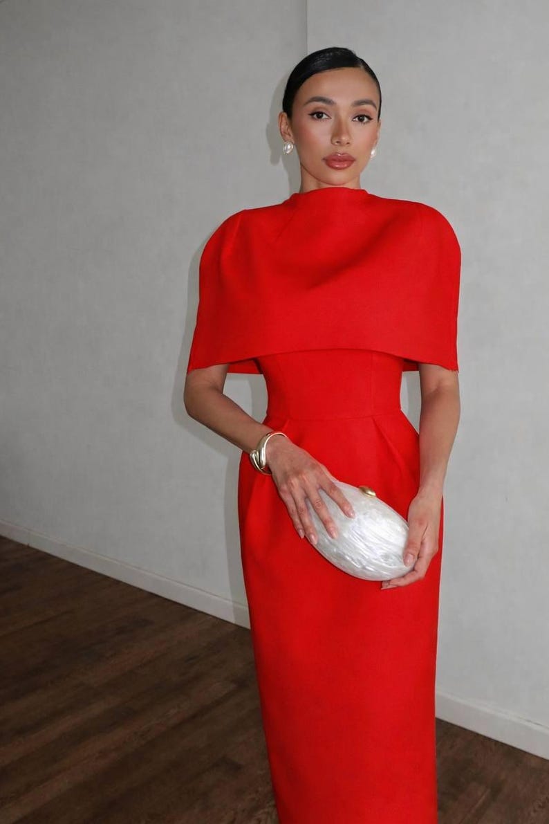 K&ouml;nnte beinhalten: Ein leuchtend rotes Kleid mit einem passenden, kapuzenartigen Oberteil. Das Kleid wird mit einer wei&szlig;en Clutch und einem goldenen Armband kombiniert. Das Outfit ist ein mutiges Statement, perfekt f&uuml;r formelle Anl&auml;sse.