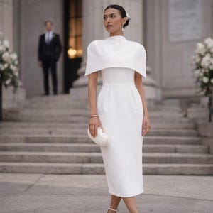 K&ouml;nnte beinhalten: Eine Frau in einem wei&szlig;en Kleid mit hohem Ausschnitt und kapuzenartigen &Auml;rmeln steht vor einem Geb&auml;ude mit gro&szlig;en wei&szlig;en Blumenarrangements. Sie h&auml;lt eine wei&szlig;e Clutch und tr&auml;gt wei&szlig;e Abs&auml;tze. Ein Mann im Anzug steht im Hintergrund.