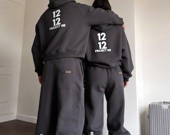 May include: Two charcoal gray hooded sweatshirts and matching sweatpants featuring the white text 12 12 PROJECT ME on the back. The sweatpants include a small back pocket and a small 12 12 logo near the ankle hem, paired with athletic sneakers.
