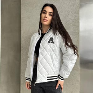 Könnte beinhalten: Weiße, gesteppte Bomberjacke mit schwarz-weißem Streifenbesatz und einem großen schwarzen Buchstaben "A" auf der Brust. Die Jacke hat ein Rautenmuster und einen Reißverschluss. Kleidung.