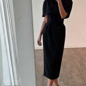 K&ouml;nnte beinhalten: Ein schwarzes Midikleid mit einem capeartigen &Uuml;berwurf an den Schultern. Das Kleid hat ein figurbetontes Oberteil und einen geraden Rock, kombiniert mit schwarzen Riemchen-High Heels. Der Gesamtstil ist elegant und f&uuml;r formelle Anl&auml;sse geeignet.