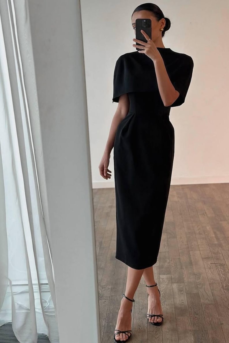 K&ouml;nnte beinhalten: Ein schwarzes Midikleid mit einem capeartigen &Uuml;berwurf an den Schultern. Das Kleid hat ein figurbetontes Oberteil und einen geraden Rock, kombiniert mit schwarzen Riemchen-High Heels. Der Gesamtstil ist elegant und f&uuml;r formelle Anl&auml;sse geeignet.