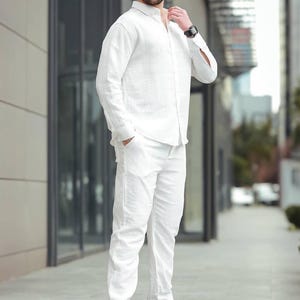 May include: A white linen shirt and trousers outfit. The ensemble features a long-sleeved button-down shirt and straight-leg trousers. The person is wearing white trainers with black detailing and sunglasses. A watch is visible on the wrist.