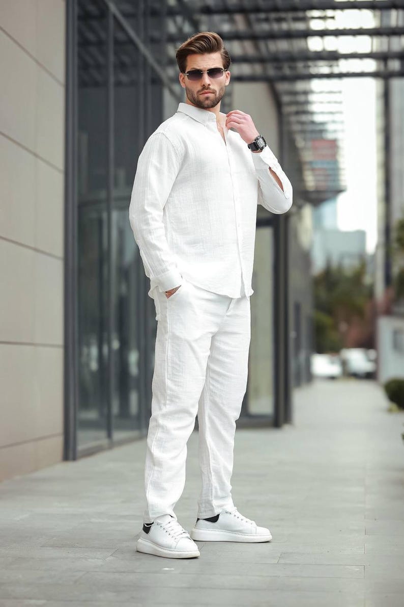 May include: A white linen shirt and trousers outfit. The ensemble features a long-sleeved button-down shirt and straight-leg trousers. The person is wearing white trainers with black detailing and sunglasses. A watch is visible on the wrist.