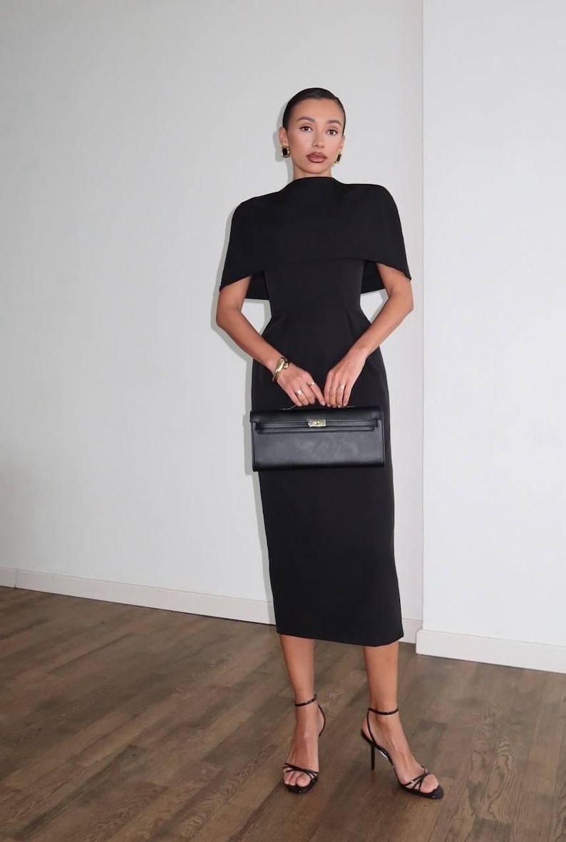 K&ouml;nnte beinhalten: Schwarzes Midikleid mit capeartigem &Uuml;berwurf, kombiniert mit schwarzen High-Heel-Sandalen und einer schwarzen Clutch. Das Kleid hat ein figurbetontes Oberteil und einen knielangen Rock. Goldschmuck akzentuiert den Look.