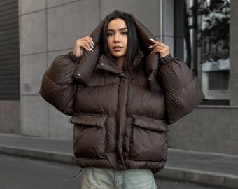 May include: A dark brown puffer jacket with a hood, long sleeves with elastic cuffs, and two large front pockets with flaps. The jacket features a front zipper closure and a quilted design, paired with light blue denim jeans.