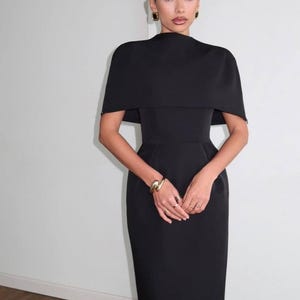 K&ouml;nnte beinhalten: Ein schwarzes Kleid mit einem kapuzenartigen &Uuml;berzug. Das Kleid hat eine figurbetonte Silhouette und eine Midi-L&auml;nge. Das Model tr&auml;gt Goldschmuck, darunter ein Armband und Ohrringe. Der Hintergrund ist eine schlichte wei&szlig;e Wand.