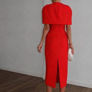 K&ouml;nnte beinhalten: Ein leuchtend rotes Kleid mit passendem cape-&auml;hnlichem Oberteil. Das Kleid hat eine figurbetonte Silhouette mit einem Schlitz hinten. Das Outfit wird mit wei&szlig;en Abs&auml;tzen und einer Clutch kombiniert. Der Look ist elegant und f&uuml;r formelle Anl&auml;sse geeignet.