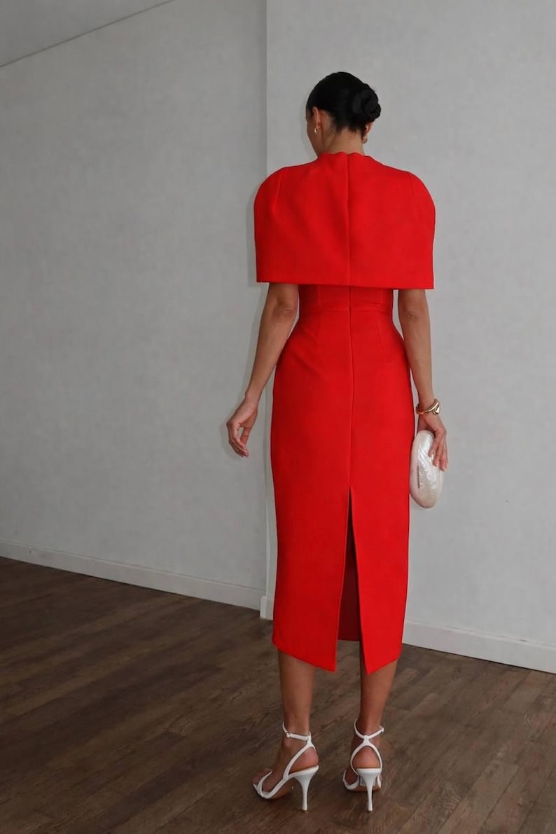 K&ouml;nnte beinhalten: Ein leuchtend rotes Kleid mit passendem cape-&auml;hnlichem Oberteil. Das Kleid hat eine figurbetonte Silhouette mit einem Schlitz hinten. Das Outfit wird mit wei&szlig;en Abs&auml;tzen und einer Clutch kombiniert. Der Look ist elegant und f&uuml;r formelle Anl&auml;sse geeignet.