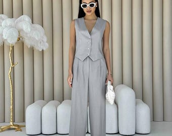 May include: A light gray vest and wide-leg pants suit. The sleeveless vest has a V-neck and button closure. The pants are high-waisted with a pleated front. A white handbag and sunglasses complete the outfit.