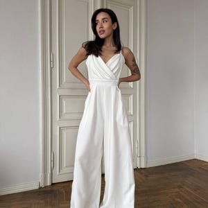 May include: A white jumpsuit with a draped bodice and wide-leg trousers. The sleeveless top features a V-neckline and spaghetti straps. The jumpsuit has a tailored fit and is made of a smooth, flowing fabric. The model is standing in front of a white door.