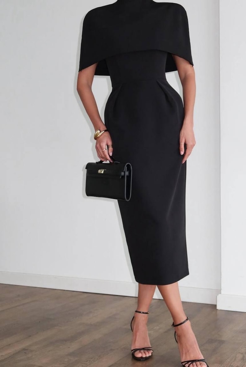 K&ouml;nnte beinhalten: Ein schwarzes Midikleid mit einem kapuzenartigen &Uuml;berzug, kombiniert mit schwarzen Riemchen-High Heels und einer kleinen schwarzen Handtasche. Das Kleid hat ein figurbetontes Oberteil und einen knielangen Rock. Ein goldenes Armband sorgt f&uuml;r einen Hauch von Eleganz.