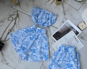 May include: A matching set consisting of a strapless tube top, a pair of shorts, and a pair of trousers, all featuring a blue and white floral toile de jouy pattern. The garments have elasticized waistbands and gathered details throughout the fabric.