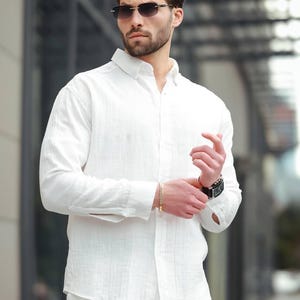 May include: A white linen shirt and matching trousers are worn with a gold bracelet and a black watch. The person is also wearing dark-framed sunglasses. The outfit is a casual, yet stylish ensemble.