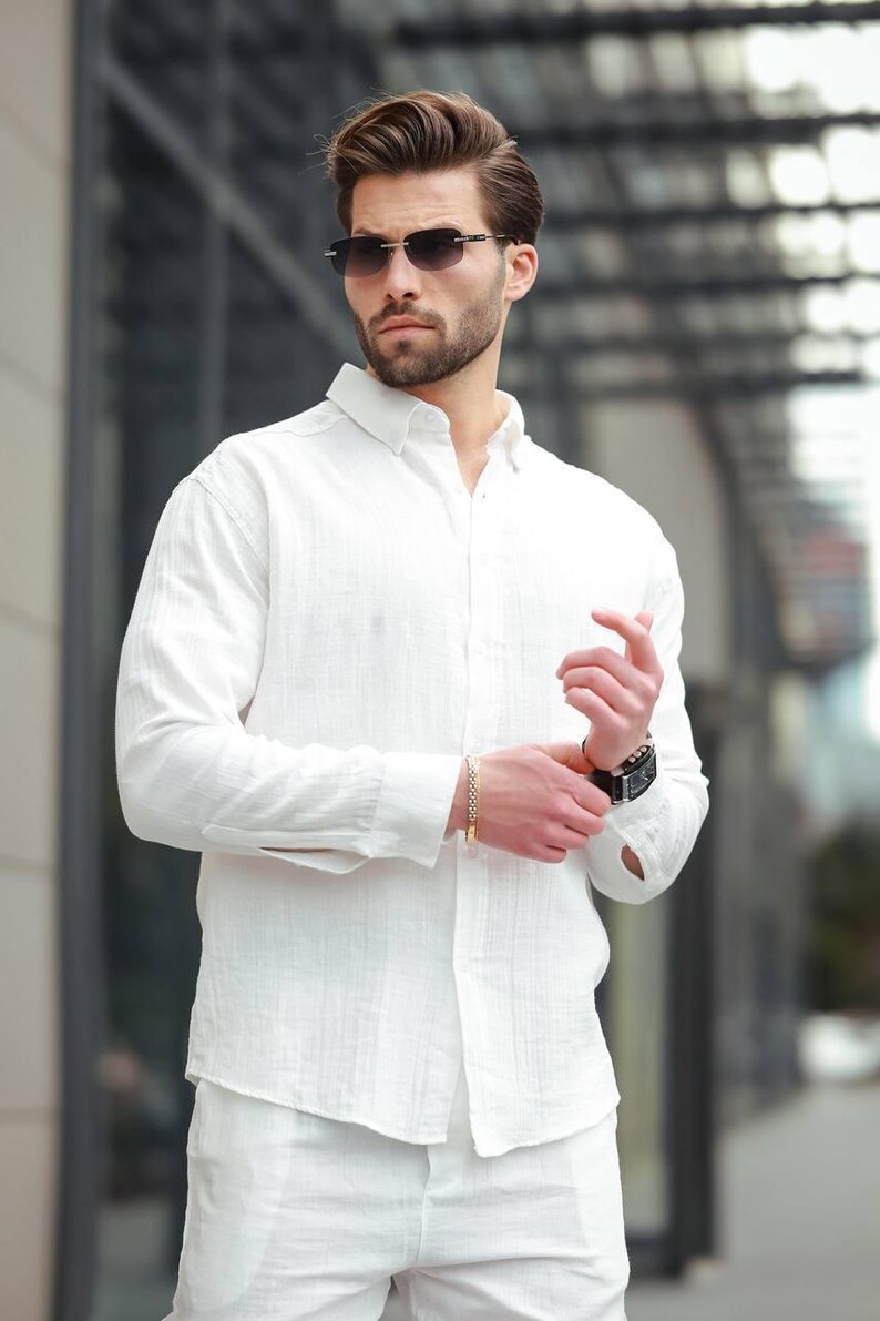 May include: A white linen shirt and matching trousers are worn with a gold bracelet and a black watch. The person is also wearing dark-framed sunglasses. The outfit is a casual, yet stylish ensemble.