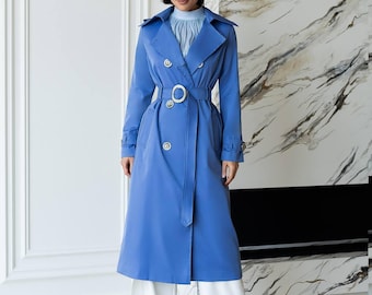 May include: A long, bright blue trench coat featuring a double-breasted front with metallic buttons, a matching waist belt with a circular buckle, and wide lapels. The coat is worn over a light blue pleated high-neck top and wide-leg white trousers.