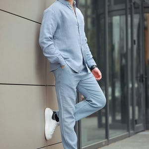 May include: A light blue linen shirt and trousers co-ord. The long-sleeved shirt is buttoned up and paired with straight-leg trousers. The outfit is completed with white trainers. The person is wearing sunglasses.