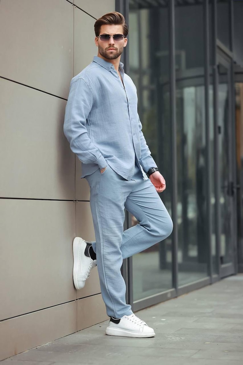 May include: A light blue linen shirt and trousers co-ord. The long-sleeved shirt is buttoned up and paired with straight-leg trousers. The outfit is completed with white trainers. The person is wearing sunglasses.
