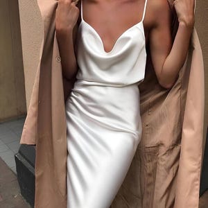 Puede incluir: Un vestido lencero de sat&eacute;n blanco con cuello desbocado y tirantes finos. Una gabardina beige est&aacute; drapeada sobre los hombros. El vestido tiene largo midi y un ligero brillo.