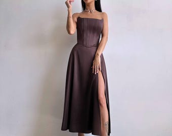 May include: A dark brown midi dress featuring a strapless corset-style bodice with vertical seams and a long, flowing skirt with a high side slit. The garment is made of a smooth, satin-like fabric and is paired with black open-toe slide sandals.