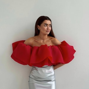 May include: A red, off-the-shoulder top with a ruffled design and a silver mini skirt. The top has a voluminous, layered ruffle, creating a dramatic effect. The skirt has a shiny, metallic finish.