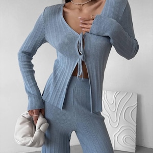 May include: A light blue ribbed knit cardigan and matching wide-leg pants set. The cardigan has a V-neck and tie closure. A white, knotted clutch bag is held in the hand. The outfit is a stylish ensemble.