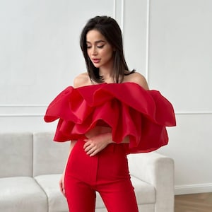 May include: A vibrant red off-the-shoulder top with a layered, ruffled design paired with matching red trousers. The outfit is a bold, monochromatic ensemble, perfect for a statement look. The top's voluminous ruffles add a dramatic flair to the outfit.