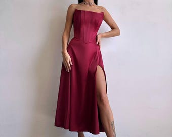 May include: A strapless burgundy midi dress featuring a structured corset-style bodice with vertical seams and a flowing skirt with a high side slit. The garment is made of a smooth, satin-like fabric with a subtle sheen, ending at mid-calf length.
