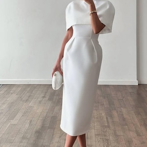 K&ouml;nnte beinhalten: Ein wei&szlig;es Midikleid mit hohem Ausschnitt und einem capeartigen &Uuml;berwurf. Das Kleid hat ein figurbetontes Oberteil und einen knielangen Rock. Das Model tr&auml;gt wei&szlig;e Riemchenabs&auml;tze und eine wei&szlig;e Clutch. Der Hintergrund ist eine wei&szlig;e Wand und ein Holzboden.
