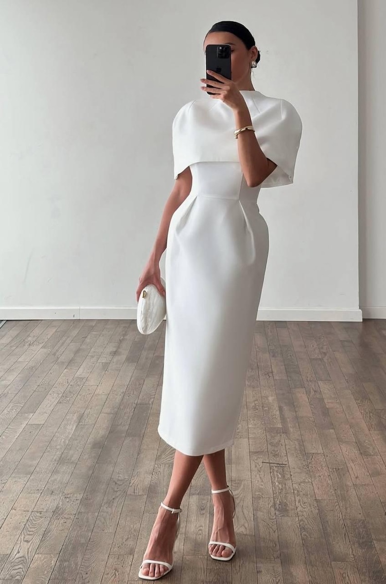K&ouml;nnte beinhalten: Ein wei&szlig;es Midikleid mit hohem Ausschnitt und einem capeartigen &Uuml;berwurf. Das Kleid hat ein figurbetontes Oberteil und einen knielangen Rock. Das Model tr&auml;gt wei&szlig;e Riemchenabs&auml;tze und eine wei&szlig;e Clutch. Der Hintergrund ist eine wei&szlig;e Wand und ein Holzboden.