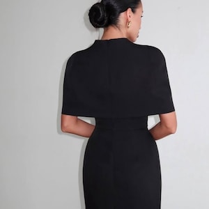 K&ouml;nnte beinhalten: Ein schwarzes Kleid mit einem kapuzenartigen Oberteil und einem figurbetonten Rock. Das Kleid hat einen hohen Ausschnitt und kurze &Auml;rmel. Das Haar des Models ist zu einem Dutt gestylt, und sie tr&auml;gt goldene Creolen. Das Kleid ist ein klassisches und elegantes Design.