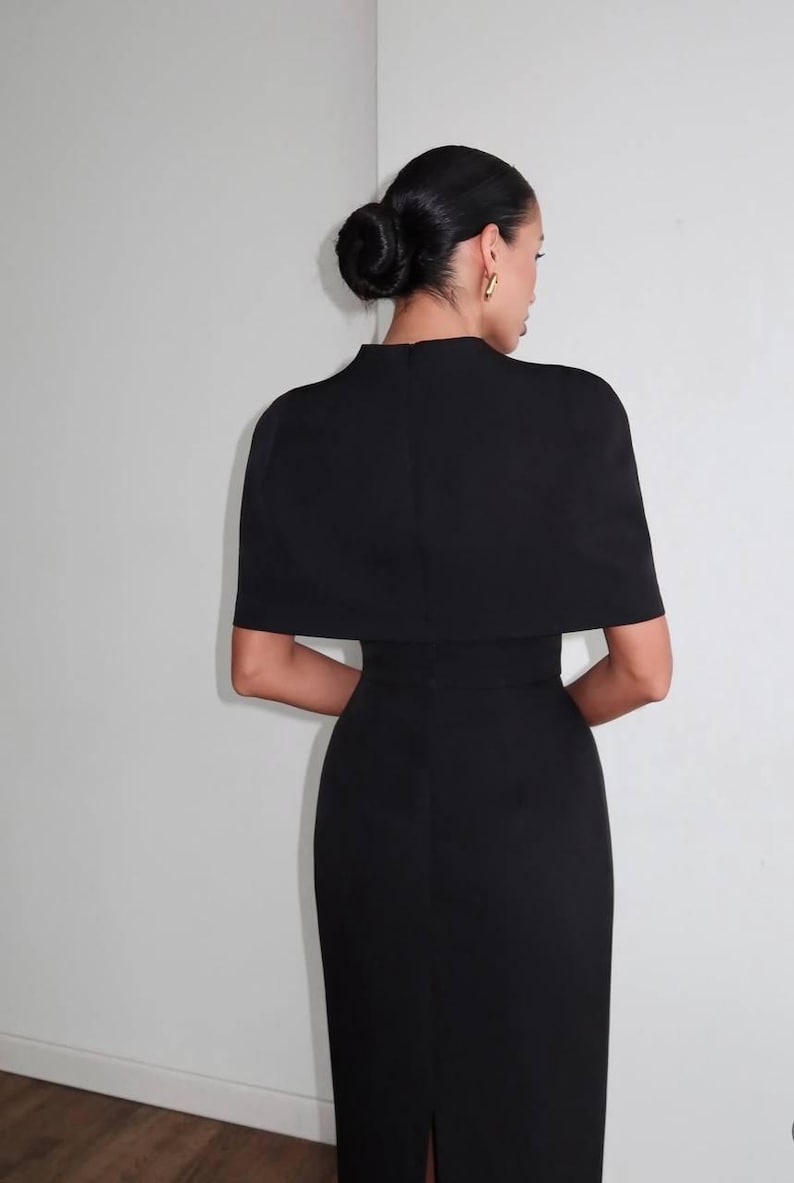 K&ouml;nnte beinhalten: Ein schwarzes Kleid mit einem kapuzenartigen Oberteil und einem figurbetonten Rock. Das Kleid hat einen hohen Ausschnitt und kurze &Auml;rmel. Das Haar des Models ist zu einem Dutt gestylt, und sie tr&auml;gt goldene Creolen. Das Kleid ist ein klassisches und elegantes Design.