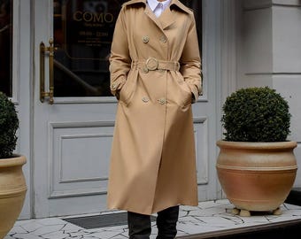 May include: A beige double-breasted trench coat featuring a wide collar, long sleeves with buttoned cuffs, and a matching waist belt with a circular buckle. The coat is worn over a white collared shirt and paired with dark gray knee-high slouchy boots.