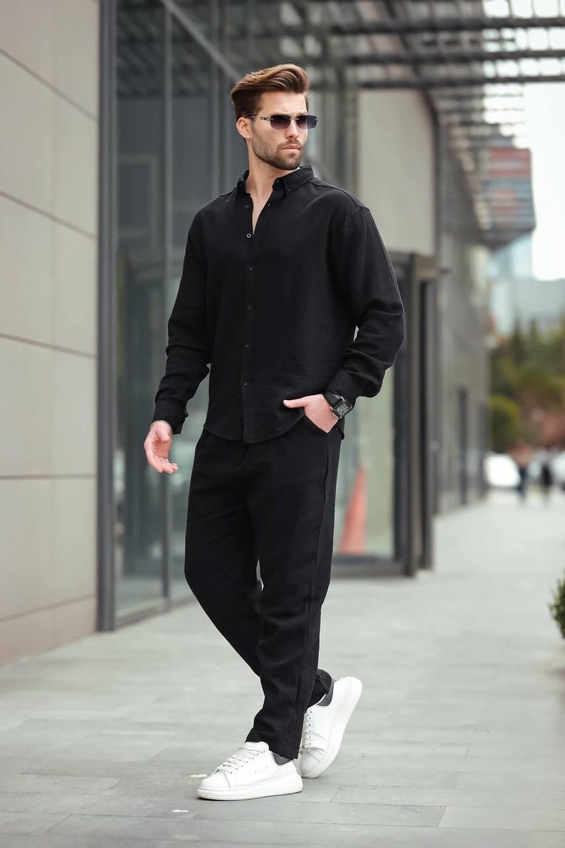 May include: A person in a black long-sleeved shirt and trousers, paired with white trainers and sunglasses. The outfit is a coordinated, all-black ensemble. The look is suitable for various occasions, from casual to smart-casual.