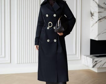 May include: A long black double-breasted trench coat featuring large silver buttons, a matching belt with a circular silver buckle, and epaulets on the shoulders. The coat is worn over a dark brown top and skirt, paired with dark tights and pointed heels.