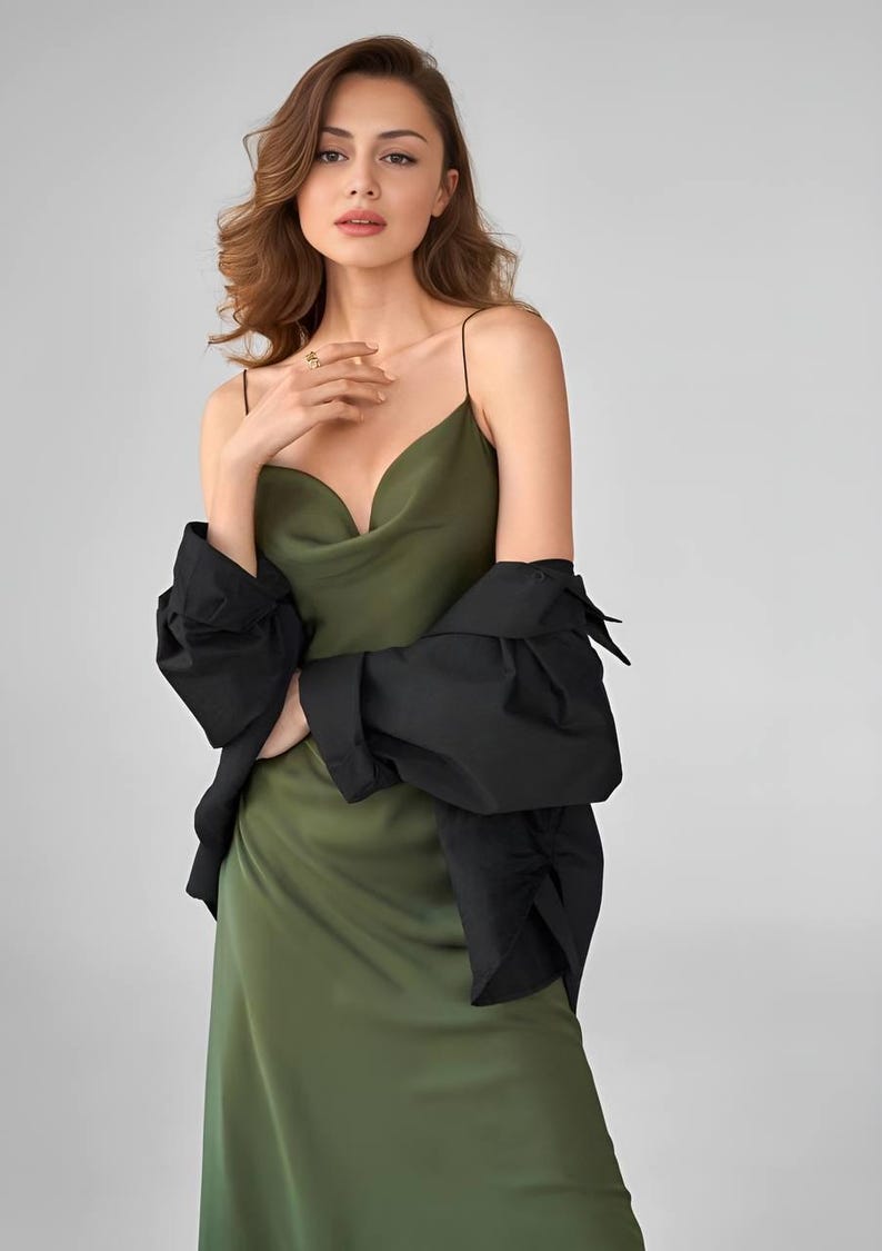 Puede incluir: Un vestido lencero verde oliva con tirantes finos y cuello desbocado. Una chaqueta negra est&aacute; drapeada sobre los hombros. El vestido es de un material sedoso. La modelo lleva un anillo dorado.