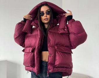 May include: A deep burgundy puffer jacket with a large hood, long sleeves, and two front flap pockets. The jacket features a front zipper closure with snap buttons, a high collar, and an adjustable drawstring hem, worn over a black crop top and jeans.